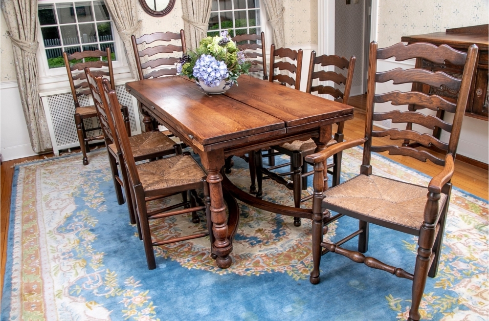A Louis XIII style antique French oak hunt table with two drop down leaves rests on turned legs conjoined with two c-form stretchers and a set of eight English oak Windsor chairs all upon a beautiful bright blue Persian rug.